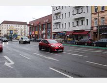 Straßenszene in einer urbanen Umgebung mit mehrspuriger Fahrbahn, auf der Autos fahren. Am Straßenrand befinden sich Geschäfte in einem roten Klinkergebäude sowie Wohnhäuser, darunter ein U-Bahn-Eingang mit dem Schild „U Hermannstraße“.
