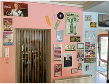 Innenraum mit rosa und hellblauen Wänden, dekoriert mit zahlreichen Retro-Blechschildern und Plaketten (u. a. Getränke- und Diner-Motive). In der Mitte befindet sich eine Türöffnung mit Lamellenvorhang, darunter ein Heizkörper; rechts führt ein Glasdurchgang zu einem hellen Wintergartenbereich.