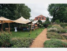 Garten mit grünem Rasen, bepflanzten Beeten und einer mit Kies bestreuten Wegfläche; mehrere große, beige Zelte mit Sitzgelegenheiten und ein Tischkicker befinden sich unter schattigen Bäumen, während im Hintergrund ein Gebäude mit rotem Schrägdach und symmetrischen Fenstern steht.