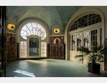 Jugendstil-Innenraum mit türkisfarbener Decke, verzierten Wandleuchten und großen Rundbogenfenstern mit kunstvollen Verglasungen. Im Vordergrund steht eine Topfpflanze, die auf ein einladendes Ambiente hinweist.