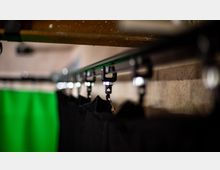 Nahaufnahme einer Metallstange mit daran befestigten schwarzen Haken, die dunkle Stoffvorhänge halten. Im Hintergrund ist ein unscharfer, grüner Vorhang zu sehen.