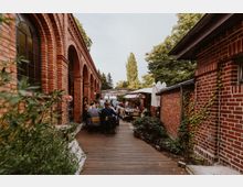 Ein Innenhof mit rotem Backstein, Arkaden an der linken Seite und Tischen, an denen Menschen sitzen. Rechts sind weitere Backsteinwände mit Pflanzen, während im Hintergrund Bäume und Sonnenschirme sichtbar sind.