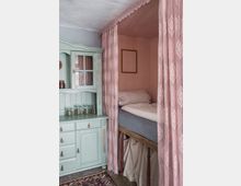 Ein rustikaler Innenraum mit einem mintgrünen Holzschrank auf der linken Seite und einem eingebauten Bett, das durch rosa Vorhänge mit Spitzenmuster abgetrennt ist. Im Vordergrund liegt ein Teppich, und im Bettbereich sind einfache Bettlaken platziert.