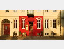 Fassade eines historischen Gebäudes mit rotem Eingangsbereich, beschriftet mit „Ballhaus Wedding“. Der Eingangsbereich hat eine geschwungene Markise, flankiert von großen weißen Fenstern mit roten Vorhängen, und darüber einen kleinen Balkon.