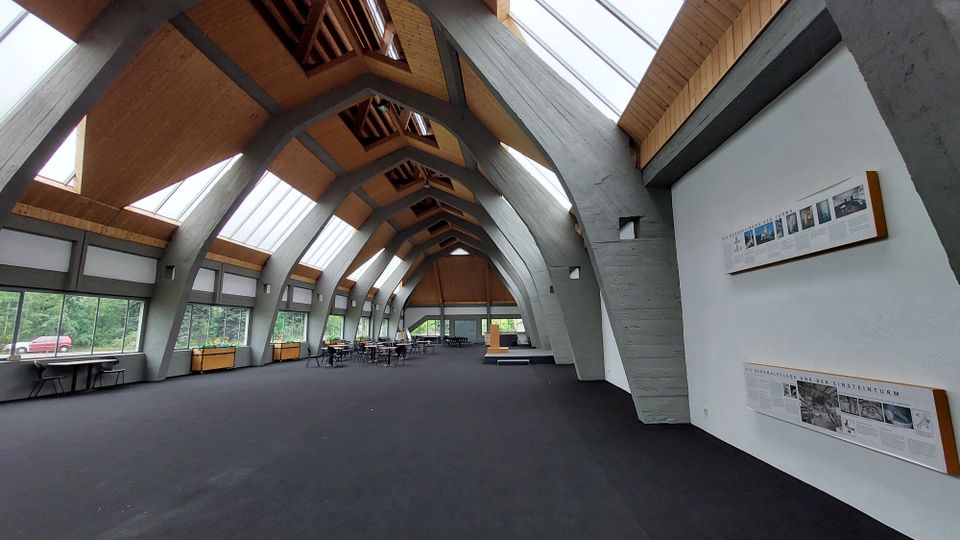 Großer Innenraum mit moderner Architektur, geprägt von einer gewölbten Holz- und Betondecke mit großzügigen Fensterfronten, die viel Tageslicht einlassen. An den Wänden befinden sich Informationstafeln, und im Raum stehen Tische und Stühle.