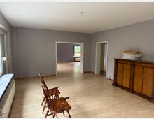 Wohnraum mit hellem Laminatboden, grauen Wänden und großen Fenstern auf der linken Seite. Im Vordergrund befinden sich zwei Holzstühle, rechts ein antikes Holzsideboard; im Hintergrund sind weitere Räume sichtbar.