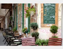 Eine enge Gasse mit draußen stehenden Tischen und Stühlen eines Cafés vor einer rustikalen Fassade mit grünen Fensterläden und Blumentöpfen. Eine Kreidetafel neben der Tür zeigt das Menü, während Fahrräder entlang der Wand geparkt sind.