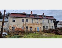 Mehrstöckiges, heruntergekommenes Gebäude mit bröckelnder Fassade und sichtbar freigelegtem Mauerwerk. Im Vordergrund ungemähte Vegetation, links ein abgestelltes Fahrzeug und ein gestutzter Baumstumpf auf der rechten Seite.