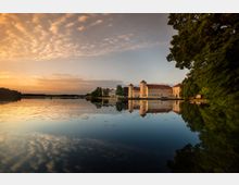 Schloss am Wasser mit zwei Türmen, dessen helle Fassade in einem ruhigen See gespiegelt wird. Der See wird von Bäumen am Ufer und einem warmen, orangefarbenen Sonnenuntergang im Hintergrund umrahmt.