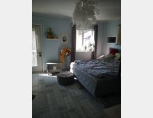 Ein Schlafzimmer mit hellblauen Wänden und Holzboden in grauer Optik. Es enthält ein Doppelbett mit grauer Bettwäsche, ein großes Fenster mit Gardinen, eine Hängelampe aus dekorativen weißen Elementen und eine Tür, die auf einen Balkon führt.