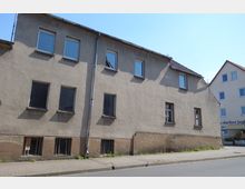 Zweistöckiges Wohngebäude mit einfacher Putzfassade, mehreren rechteckigen Fenstern und einem schmalen Gehweg davor. Das Gebäude wirkt älter, mit einigen kleineren Schäden an der Fassade, nebenan ist ein moderneres Gebäude erkennbar.