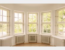 Ein heller Raum mit einer halbrunden Fensterfront, bestehend aus fünf großen Sprossenfenstern. Unter jedem Fenster befinden sich weiße Heizkörper, und durch die Fenster ist Grünfläche mit Bäumen sichtbar.