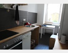 Kleine Küche mit rechteckiger Arbeitsfläche aus Holz, Spüle, Herd und Geschirrspüler. Die Rückwand ist mit dunkelgrauen Fliesen verkleidet, ein Fenster lässt Tageslicht in den Raum, und im Hintergrund steht ein kleiner weißer Tisch.