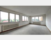 Leerstehender Raum mit weiß gestrichenen Wänden, großen rechteckigen Fenstern an zwei Seiten und grauem Boden. Unter den Fenstern befinden sich weiße Heizkörper, und durch die Fenster ist eine Aussicht auf Dächer und Bäume sichtbar.