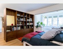 Schlafzimmer mit großer Holzbücherwand, einem Doppelbett mit blauem Bettbezug und rotem Überwurf sowie einer angrenzenden hellen Nische mit großen Fenstern, durch die Tageslicht einfällt. In der Nische befinden sich eine Badewanne und Zimmerpflanzen.