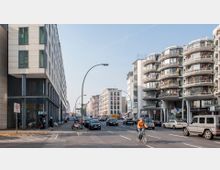 Stadtszene mit einer breiten Straße, flankiert von modernen Gebäuden. Auf der linken Seite ein Hotel mit glatter Fassade, auf der rechten Seite Wohnhäuser mit runden Balkonen; Autos und ein Fahrradfahrer sind auf der Straße zu sehen.