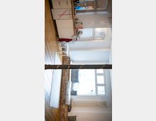 Heller Wohnraum mit offener Küchenzeile links, Holzfußboden und weiß verputzten sowie teilweise freiliegenden Ziegelwänden. Vor großen Fenstern befindet sich eine gepolsterte Sitzbank auf gemauerter Basis, in der Mitte steht eine dunkle Stütze; ein Teppich und ein kleiner Beistelltisch ergänzen den Raum.