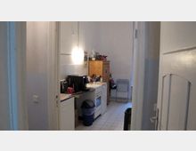 Kleine, schmale Küche mit weißen Fliesen und heller Wandfarbe. Zu sehen sind eine Arbeitsplatte mit Mikrowelle, ein Herd, ein Holzschrank, ein Mülleimer und ein Heizkörper mit einem Abtropfgestell davor.