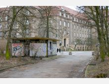 Eine verschmutzte und mit Graffiti bedeckte kleine Hütte steht auf einem unbefestigten Weg. Im Hintergrund sieht man ein großes historisches Gebäude mit symmetrischen Fenstern, einem Ziegeldach und einer verzierten Eingangstür, umgeben von kahlen Bäumen in einer urbanen Umgebung.