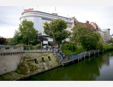 Ein Stadtbild mit einem Fluss im Vordergrund, einer steinernen Ufermauer mit Moosbewuchs und einer Promenade. Im Hintergrund ist ein modernes Gebäude mit einer geschwungenen Glasfassade und Aufschrift „Domäne“ sowie ein historisches Backsteingebäude mit Giebeln zu sehen, umgeben von Bäumen und Vegetation.