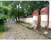 Pflasterweg entlang einer weiß verputzten Mauer mit Verzierungen aus roten Ziegeln und einem Holztor. Auf der rechten Seite steht ein großer Baum vor der Mauer, im Vordergrund ein Blumentopf mit weißen Blumen, und links im Hintergrund sind geparkte Autos und Boote zu sehen.