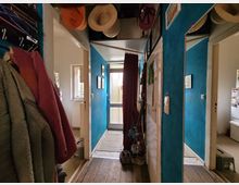 Schmaler Flur in einer Wohnung mit Holzboden und blau gestrichenen Wänden. Links hängen Mäntel, Hüte und Taschen an Haken, rechts ist ein großer Spiegel sichtbar, und im Hintergrund führt eine verglaste Tür nach draußen.