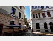 Straßenszene mit einem Café auf der linken Seite, das durch Schriftzug „Kaffeestube“ gekennzeichnet ist, und einem angrenzenden Gebäude mit dekorativen weißen Fassaden und Rundbogenfenstern. Vor dem Café stehen Sonnenschirme und Tische auf gepflastertem Boden, im Hintergrund Bäume und Laternen.