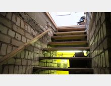 Holztreppe in einem schmalen Kelleraufgang mit Backsteinwänden, die teils roh und unverputzt sind. Unten leuchtet gelbes Licht aus einem angrenzenden Raum, und oben ist ein Teil eines eher modern eingerichteten Raumes erkennbar.