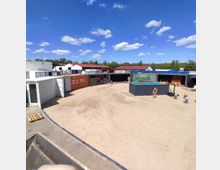 Ein offener Platz mit hellem Sandboden, umgeben von mehreren schiffscontainerartigen Strukturen in verschiedenen Farben, darunter Weiß, Orange, Grün und Blau. Im Hintergrund sind Bäume sowie flache Gebäude mit roten Dächern zu sehen, und der Himmel ist klar mit einigen Wolken.