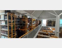 Innenansicht einer Bibliothek mit mehreren Etagen. Holzregale voller Bücher und Aktenordner stehen dicht nebeneinander, dazwischen befinden sich Tische mit Stühlen. Das Dach ist weiß und gewölbt, mit sichtbaren Stahlträgern.