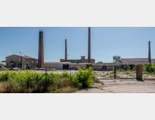 Ehemaliges Industriegebiet mit mehreren Schornsteinen aus Ziegelsteinen, umgeben von leerstehenden Gebäuden und einem verlassenen, bewachsenen Hof. Im Vordergrund ein Drahtzaun mit einem Eingangstor aus Metall und Backsteinpfeilern.