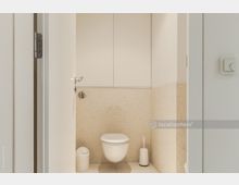 Kleines, modernes Badezimmer mit beigefarbenen Fliesen an der Wand und einem wandmontierten WC. Unterhalb von weißen Schränken befinden sich ein Mülleimer und eine Toilettenbürste.