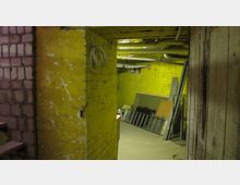 Ein einfacher Kellerraum mit gelb gestrichenen Backsteinwänden, sichtbaren Rohren an der Decke und lagernden Materialien wie Metallgittern und Holzplatten an einer Wand. Links führt eine Treppe aus dem Keller heraus.