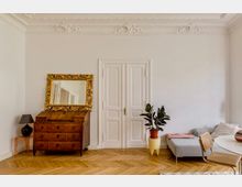 Heller Wohnraum mit weißen Wänden, Stuckdecke und Doppelflügeltür in der Mitte. Links stehen eine antike Holzkommode mit großem goldgerahmtem Spiegel und eine kleine Lampe, rechts ein graues Sofa neben einer Topfpflanze auf Fischgrätparkett.