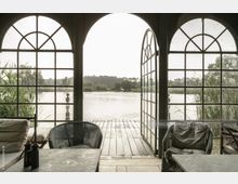 Innenansicht eines Pavillons mit drei großen, halbkreisförmig verglasten Türen, die zu einer Holzterrasse und einem See führen. Im Vordergrund stehen Sitzmöbel und ein Tisch, außen sind Schilf und Bäume sichtbar.