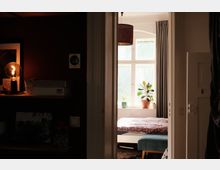 Blick durch einen Türrahmen in ein Schlafzimmer mit großem Sprossenfenster, grauen Vorhängen und Pflanzen auf der Fensterbank. Im Raum stehen ein Bett mit Decke sowie eine gepolsterte Sitzbank; links ist ein dunkler Flurbereich mit Regal und beleuchteter Lampe zu sehen.
