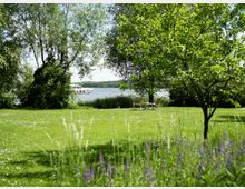 Grüne Wiese mit Bäumen und blühendem Gras im Vordergrund, dahinter zwei Holzstühle und ein Tisch mit Blick auf einen See mit einem vorbeifahrenden Boot, umgeben von dichter Vegetation.