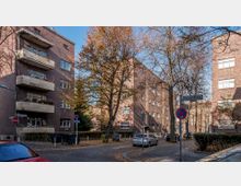 Straßenszene in einer Wohngegend mit mehrstöckigen Backsteingebäuden, Balkonen und großen Fenstern. Im Vordergrund stehen Straßenbäume mit herbstlichem Laub, geparkte Autos und ein Straßenschild mit den Namen „Elveser Straße“ und „Etzelburger Weg“.