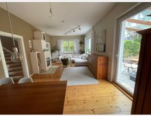 Helles Wohnzimmer mit Holzdielen, großem Ecksofa auf Teppich und einem Holzschrank mit vielen Schubladen an der Wand. Rechts führt eine bodentiefe Glasschiebetür auf eine Terrasse mit Tisch und Stühlen; links sind eine Treppe und ein moderner Kaminofen zu sehen.