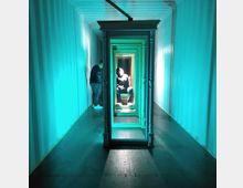 Ein schmaler, beleuchteter Raum mit türkisfarbenem Licht, der durch eine Reihe von Spiegelrahmen in der Mitte perspektivisch gestreckt wirkt. Zwei Personen befinden sich im Raum, eine Person sitzt zentral im Spiegelrahmen, während die andere seitlich steht.