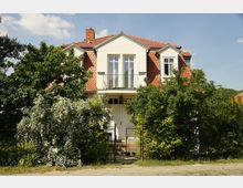 Zweistöckiges Einfamilienhaus mit cremefarbener Fassade, roten Dachziegeln und kleinen Giebelfenstern. Der Vorgarten ist dicht bepflanzt mit Sträuchern und Blüten, ein schmiedeeisernes Tor führt zur Eingangstür.