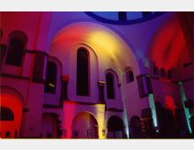 Innenraum einer Kirche mit hohen Gewölbedecken und Rundbögen, beleuchtet in bunten Farben wie Rot, Gelb und Blau. Die Fenster und architektonischen Akzente sind durch die Beleuchtung hervorgehoben.