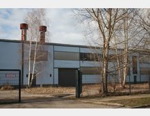 Industriegebäude mit grauen Fassaden und großen Fenstern, umgeben von kahlen Bäumen und einem Metallzaun mit einem Tor. Im Hintergrund ragen zwei rostige Schornsteine in den Himmel.