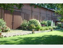 Roter Backsteinbau mit großen Holztoren, umgeben von Blumenbeeten mit Hortensien und bunten Blütenpflanzen, davor eine gepflasterte Fläche und eine Rasenfläche im Vordergrund. Ein weißer Sonnenschirm steht in der Nähe der Blumenbeete.