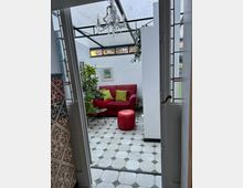 Wintergarten mit gläsernem Dach und weißen Wänden, ausgestattet mit einem roten Sofa, passenden Kissen und einem Hocker. Der Raum hat schwarz-weiße Kachelböden, Pflanzen, Kunstbilder an der Wand und einen verzierten Kronleuchter.