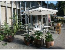 Kleiner Außenbereich eines Cafés mit weißen Tischen und Metallstühlen unter großen Sonnenschirmen. Rundherum stehen Topfpflanzen, und im Hintergrund ist ein Gebäude mit großen Fenstern zu sehen.