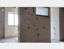 Rohbau-Interieur mit Betonwänden, sichtbaren Verfärbungen und Löchern. Im Hintergrund befindet sich ein rechteckiges Fenster mit diffusem Tageslicht, der Boden ist unbehandelt.