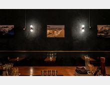 Dunkel eingerichtete Bar mit Holztischen und Stühlen im Hintergrund. An der schwarzen Wand hängen beleuchtete Bilder zwischen Wandleuchten, während im Vordergrund eine Theke mit Barutensilien zu sehen ist.