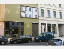 Straßenszene mit einer Reihe moderner Gebäude, darunter ein grün verputztes Haus mit großen Fenstern und einer Tür mit kreisförmigen Mustern, sowie die "Galerie Eigen + Art" im angrenzenden weißen Gebäude. Mehrere geparkte Fahrzeuge stehen entlang der Straße.