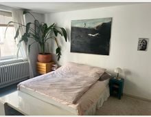 Bright bedroom with a low bed dressed in pale pink bedding against a white wall, beneath a large dark-toned painting and a small framed photo. A window with sheer curtains and a radiator sits beside a dresser topped with a large potted plant, and a small bedside table holds a lamp.